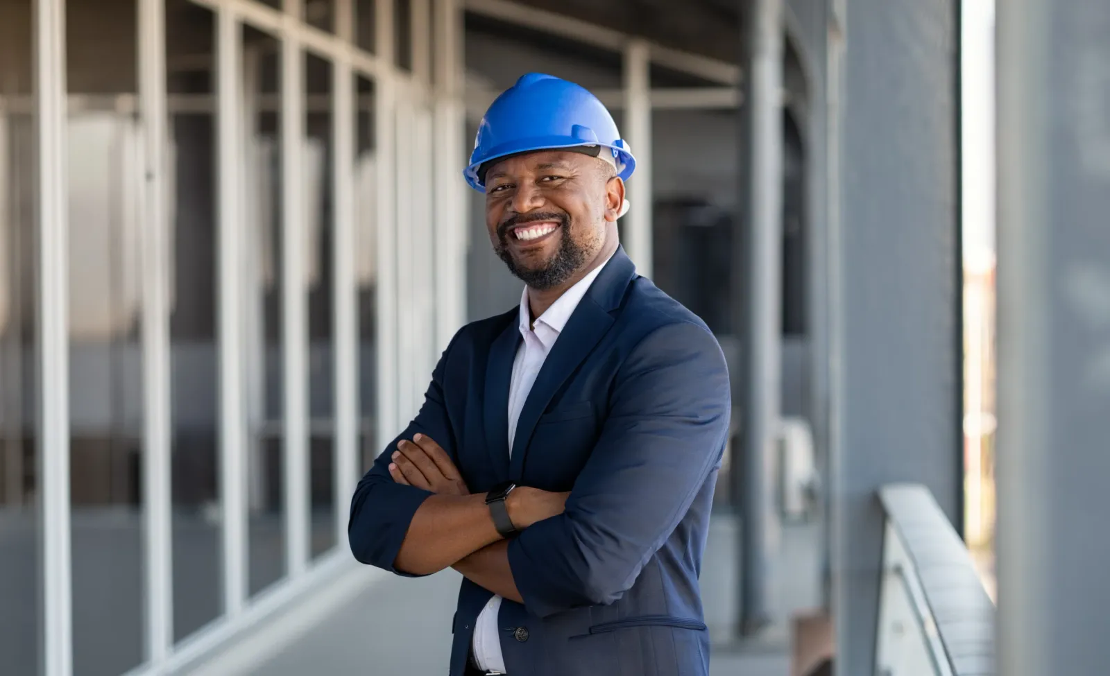 Lächelnder Bauherr mit blauem Helm und verschränkten Armen auf der Baustelle