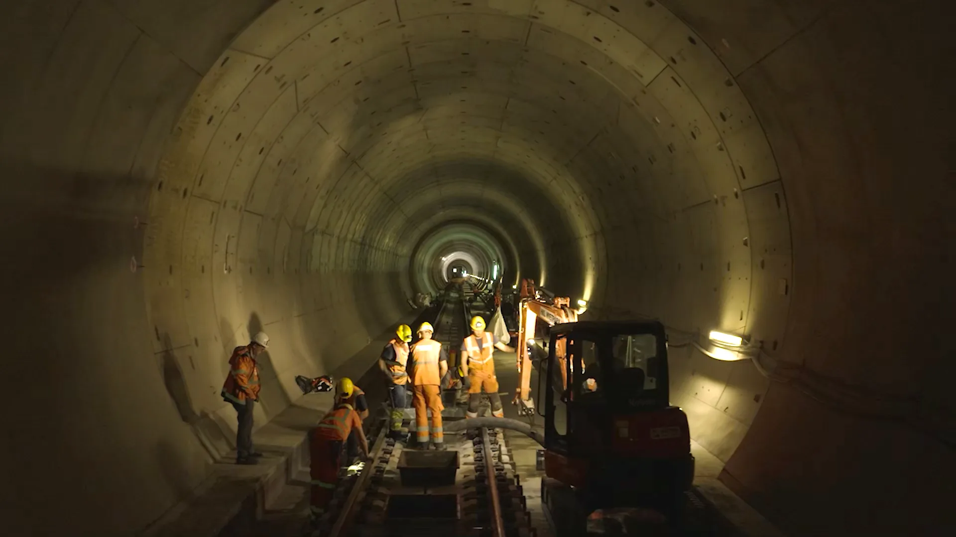 Bauarbeiter verlegen Gleise in der Tunnelröhre, im Hintergrund ein Minibagger und die beleuchtete Vortriebsstrecke